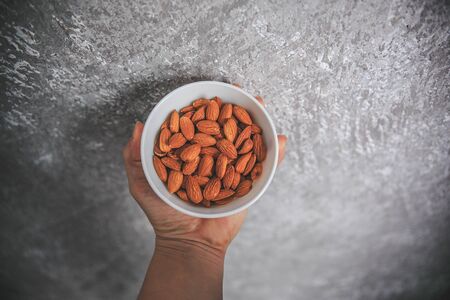 Almonds with skin without shell in the white bowl holding in hand on the gray and white background.の写真素材