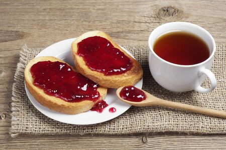 Cup of hot tea and white bread with strawberry jam in plate on old wooden tableの写真素材