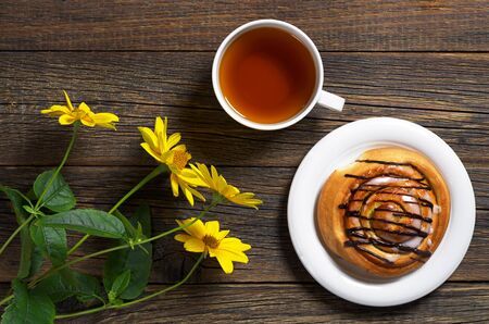 Cup of tea and sweet bun on wooden table with yellow flowers, top viewの写真素材