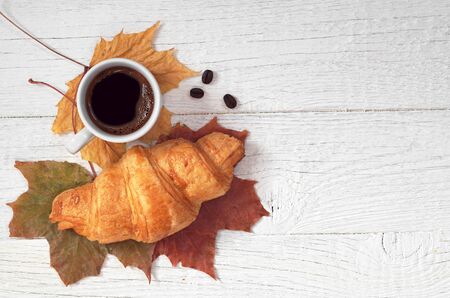 Cup of hot coffee and croissant on white wooden table with autumn leaves, top viewの写真素材