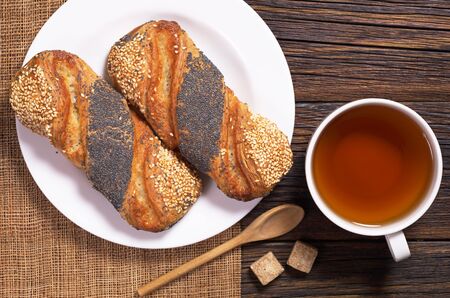 Cup of tea and buns with sesame and poppy seeds in plate on wooden table, top view