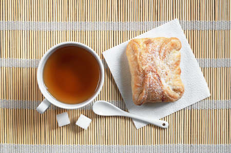 Cup of tea and sweet bun of puff pastry with jam for breakfast on a bamboo napkin background, top viewの写真素材