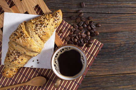 Cup of hot coffee and croissant with sesame seeds for breakfast on dark wooden table, top view