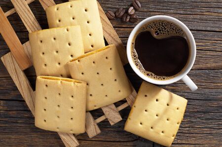 Cup of hot coffee and square cookies with jam on dark wooden table, top view