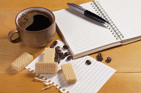 Cup of coffee with small wafers and notebook on wooden desk