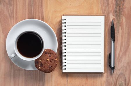 Notebook, pen and cup of coffee with chocolate cookie on wooden background. Top view の写真素材
