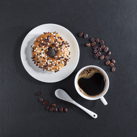 Donut decorated with white and dark chocolate balls in plate and cup of coffee on black stone background, top viewの写真素材