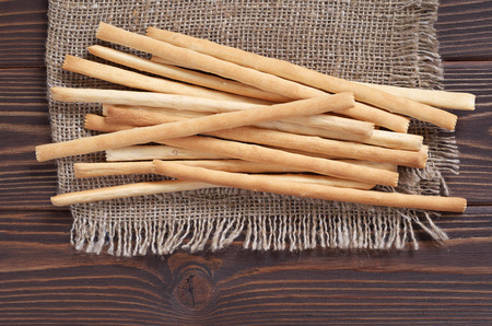 Heap of bread sticks on burlap is located on brown wooden background, top view