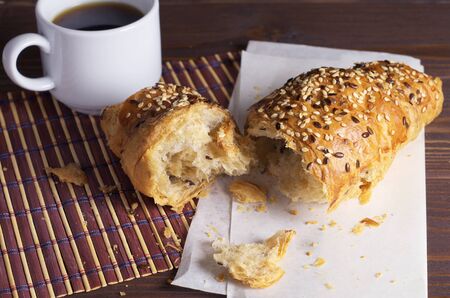 Broken croissant with seeds of flax and cup of black coffee on table close up