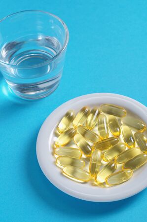 Fish oil capsules in plate and glass of water on blue background. Omega 3の写真素材