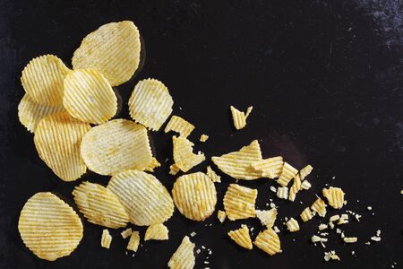 Salty ribbed potato crisps whole and broken on black tray close-up, top view with copy space
