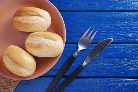 Loaves of small white bread on a plate and cutlery on blue wooden table, top view with copy space