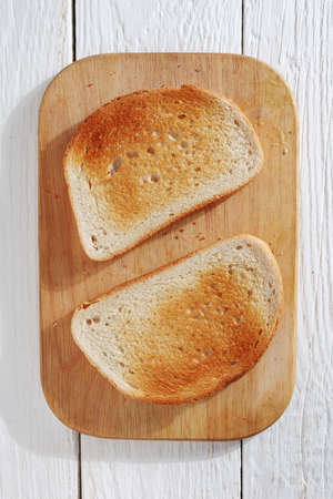Two slice of toast on cutting board on white wooden table, top view
