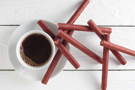 Cup of coffee and heap of chocolate wafer sticks on white wooden background, top view