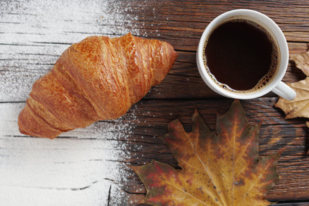 Freshly baked croissant and cup of coffee on wooden background with autumn leaves, top viewの写真素材