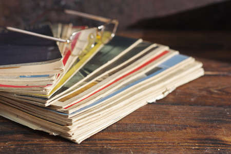 Old magazines are stacked on wooden table with reading glasses