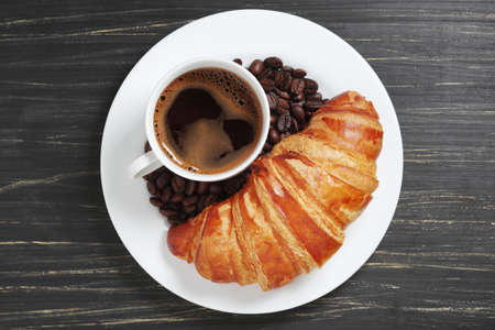 Cup of coffee and croissant on a plate on old black wooden table, top viewの写真素材