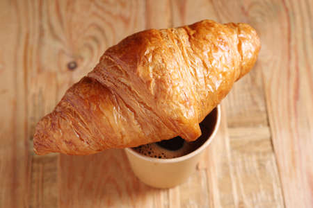 Croissant and paper disposable cup with coffee on old wooden background