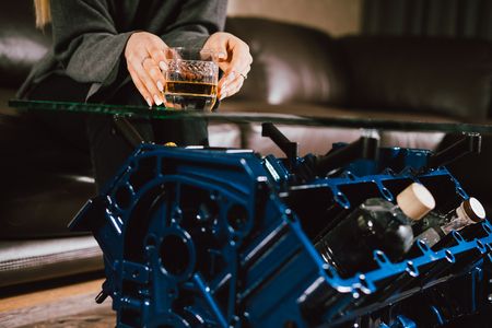 Young caucasian blonde girl sitting on sofa with glass of whiskey at luxury interior with custom v8  car engine table. Fashion picture and beautiful smileの写真素材