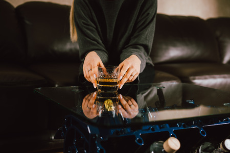 Young caucasian blonde girl sitting on sofa with glass of whiskey at luxury interior with custom v8  car engine table. Fashion picture and beautiful smileの写真素材