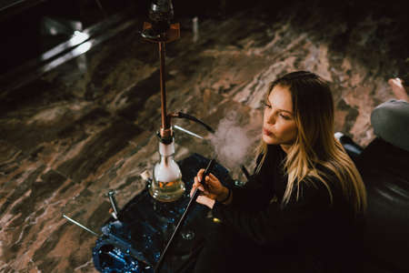 Young caucasian blonde girl smoking hookah at luxury interior with custom v8  car engine table. Leather sofa and beautiful smileの写真素材