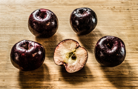 A still life of four whole crimson apples and a half of an apple laying on a wooden cutting board....