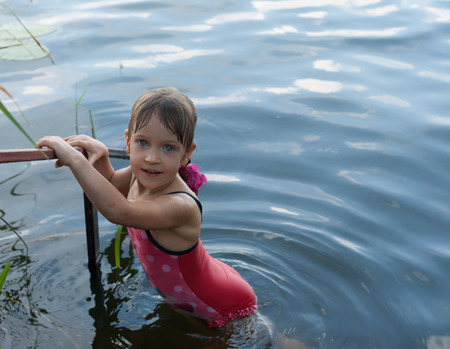 A nice four-year old girl stands holding a handrail in the open water. Child in a red swimsuit has a beautiful eyes, as blue as a water. Concept - vacation and happy childhood.の写真素材