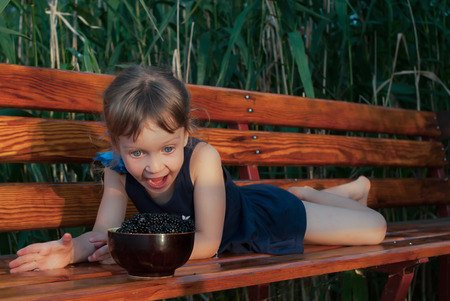 A four-years-old little girl lying on the bench among the high green grasses. The child loooks at a bowl full of fresh ripe blackberries with open mouth. Concept - expression of joy, happiness and surprise.の写真素材