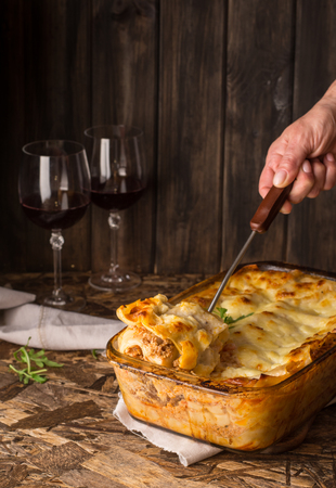 Traditional meat lasagna on wooden background. Red wine in wineglass. Homemade italian lasagna with bolognese and bechamel sause served with arugula. Gray napkin on wooden table.の写真素材