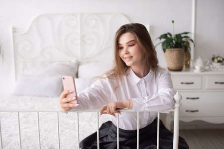 A happy European woman has a video call on her smartphone . The girl looks into the phone camera and smiles. White Scandinavian interior. Daylight through the window.の写真素材