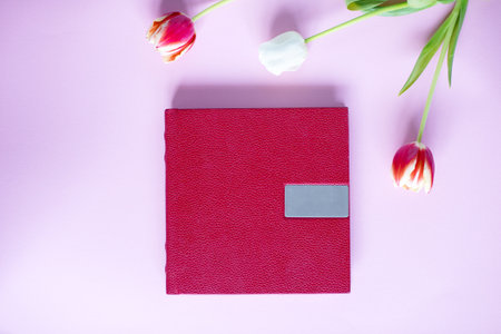 A red leather book with an embossed place for text, lies on a purple background next to flowers and tulipsの写真素材
