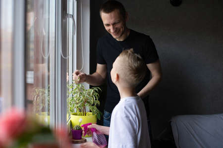 White T-shirt, dad hugs son, blonde, flower pot, watering flowers, house plants, window, window lightの写真素材