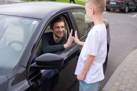 Dad smiles at his son, the child meets the father, a black car, a meeting in the city. to come home, to say goodbye near the car, to see off to work, a boy in a white T-shirt, his father's smile, an open windowの写真素材