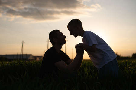 fatherhood, dads, fathers, grandfathers, adult men, family life, fatherhood, being a parent, staying at home, dads, fathers and sons, field with spikelets, green field, teenage son, blond son, sunset, black t-shirt, white t-shirt, holding each other's hand, leather belt, hold on to handsの写真素材