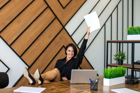 The happy woman put her feet in high-heeled shoes on the office desk and raised her hand with a...