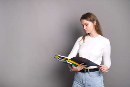 A cute student girl looks into an open folder for documents and stands on a gray background.の写真素材