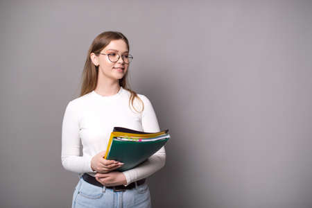 A cute student with glasses holds colorful folders on a gray background and looks awayの写真素材