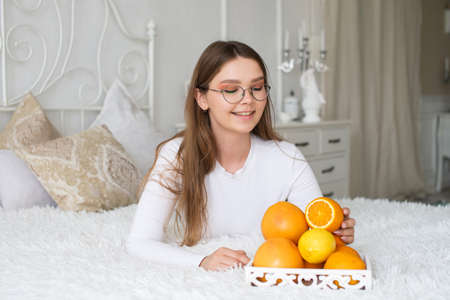 A cute girl with glasses is lying on the bed and takes part of an orange from a fruit basketの写真素材