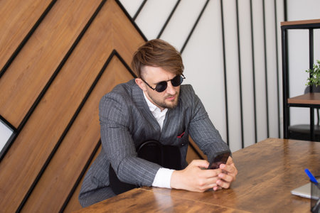 A young man in a suit and sunglasses is sitting at a desk in the office and looking at the phoneの写真素材