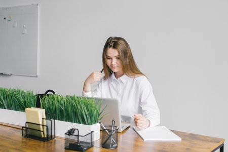 A cute girl is sitting at a wooden desk in the office working with a laptop and notepadの写真素材