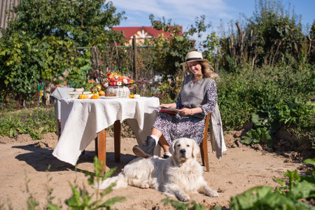 a woman in a hat is sitting on a chair near the table, a Labrador retriever is lying next to herの写真素材