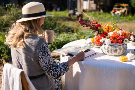 A woman in a hat with a brim flips through a book and drinks a hot drink at a table with flowers on the streetの写真素材