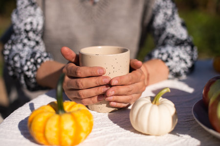 The working hands of the girl hold a ceramic cup standing on the table near small pumpkinsの写真素材