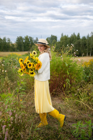 A cute girl in a brimmed hat and yellow rubber boots holds a bouquet of sunflowers in a fieldの写真素材