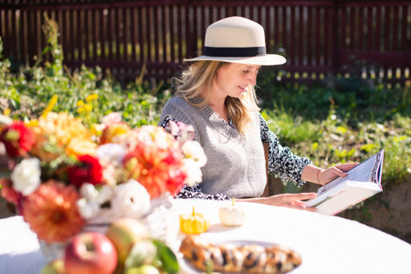 A cute girl in a hat with a brim reads a book at a table with autumn flowers on the streetの写真素材