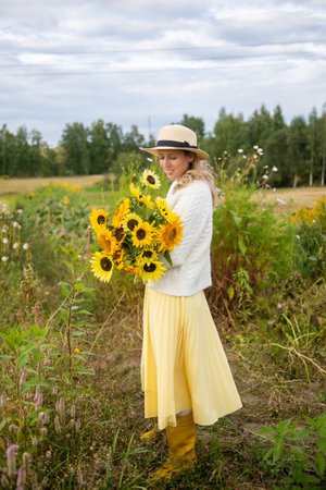 A cute girl in a brimmed hat and yellow rubber boots holds a bouquet of sunflowers in a fieldの写真素材
