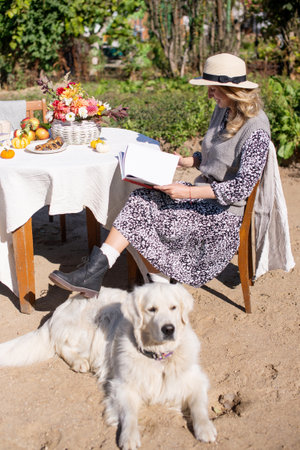a woman in a hat is sitting on a chair near the table, a Labrador retriever is lying next to herの写真素材