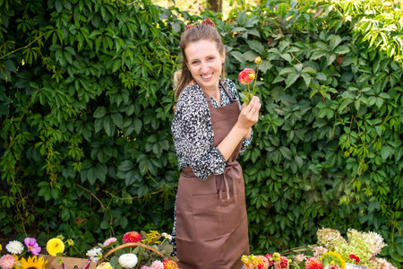 A cute girl in an apron and with a red dahlia flower is standing near a vineyardの写真素材