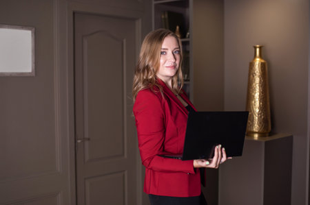 A cute girl holding an open laptop in her hands stands in the room next to the door and smilesの写真素材