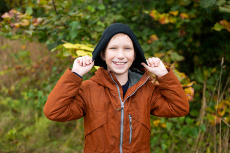 A cute boy in a red jacket puts a black hat on his head and laughsの写真素材